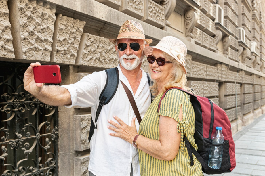 Smiling senior couple taking a selfie while traveling in Europe, enjoying retirement adventures with backpacks and summer hats.
